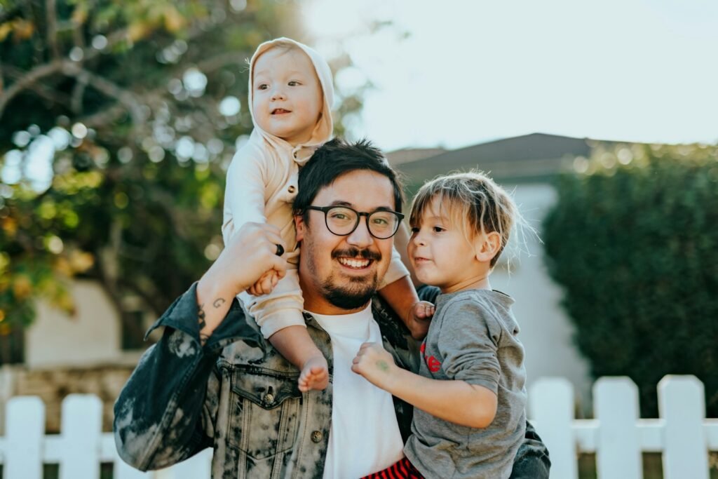 Pai sorrindo ao ar livre com dois filhos pequenos no colo e nos ombros